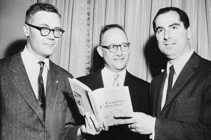 Black and white photo of three men in suits, including a young Philip Roth all holding a copy of the book Goodbye, Columbus