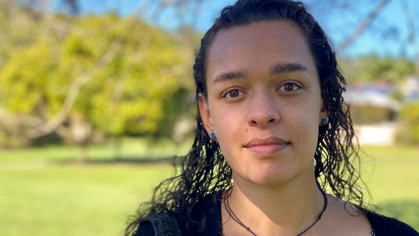 A woman in her early 20s with brown curly hair and brown eyes looks directly to the camera.