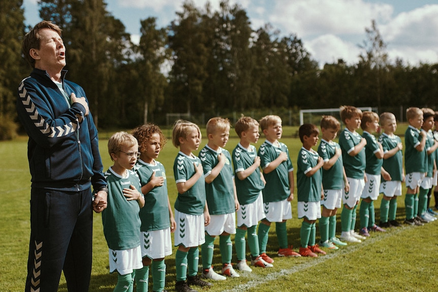 Middle-aged skinny man standing at end of row of small children in sports uniforms, all with right hands on hearts and singing.