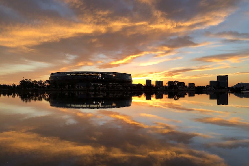 A wide shot looking over the Swan River showing Perth Stadium at dawn with clouds reflected on the water.