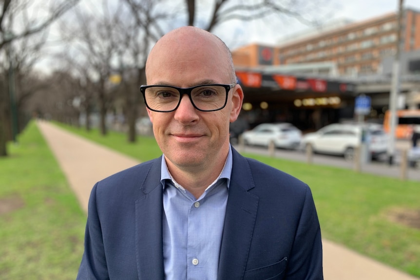 A man in a business shirt, suit jacket and glasses poses outside a hospital.