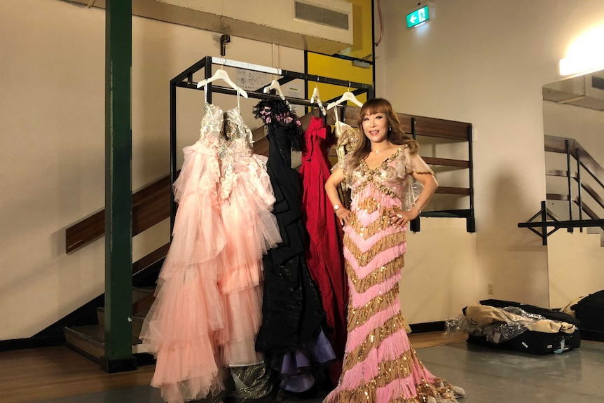 A woman in an elaborate pink and gold gown stands with other dresses on hangers