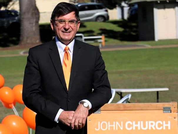 A man in a dark suit, white shirt and bright orange sign leans on a sign bearing his name.