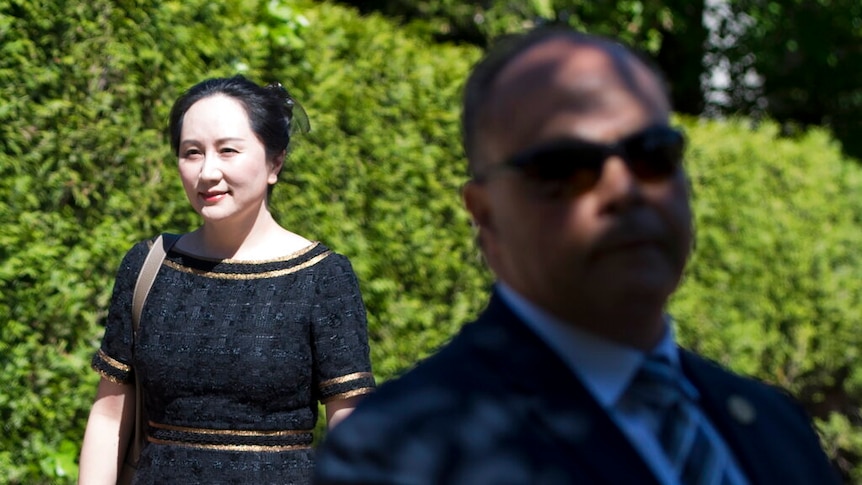 Looking past a blurred security guard, you view Meng Wanzhou in a black dress walking in front of a verdant green hedge.