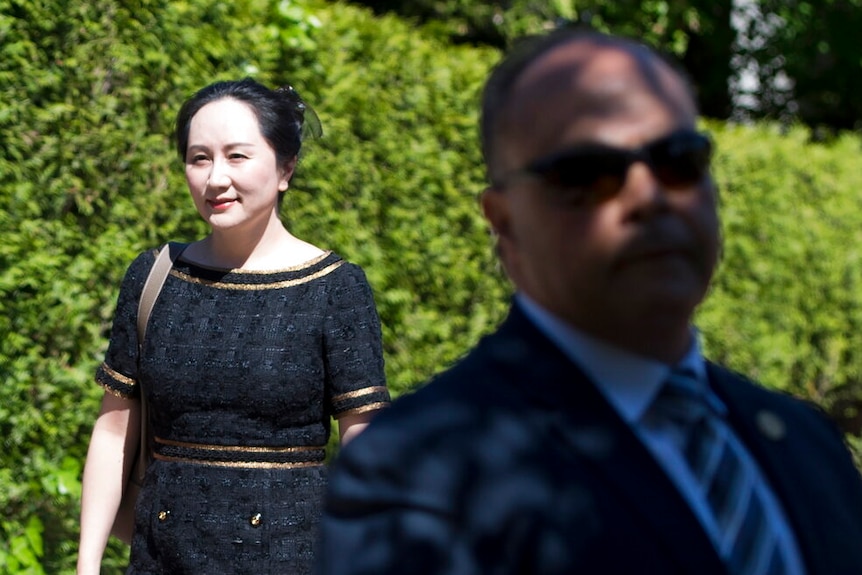 Looking past a blurred security guard, you view Meng Wanzhou in a black dress walking in front of a verdant green hedge.