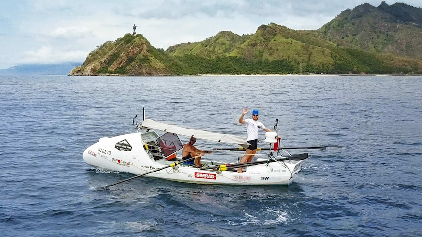 Two men in a row boat with one standing up and waving in the ocean as they row past an island.