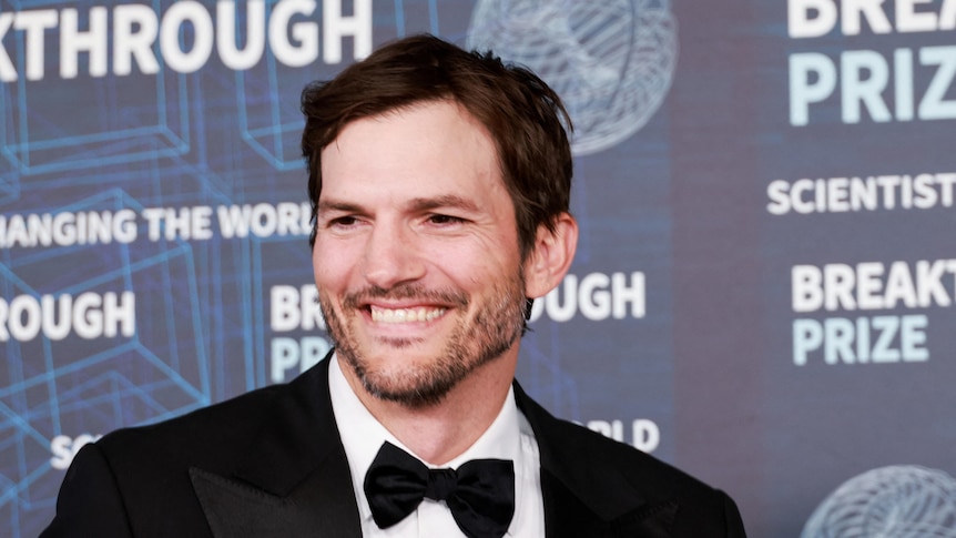 Ashton Kutcher smiles wearing a suit in front of a screen with writing on it.
