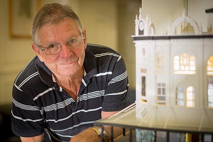 A man leans against a shelf holding a miniature model of an early 20th century building.