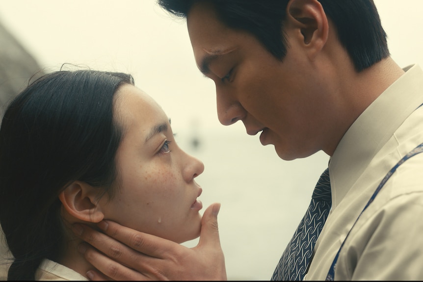 A Korean couple face each other, the man cradles the woman's face in one hand as a prelude to a kiss.