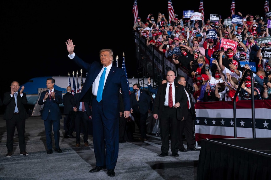 A man in a navy suit waves with his right hand while supporters in stadium seating look on.