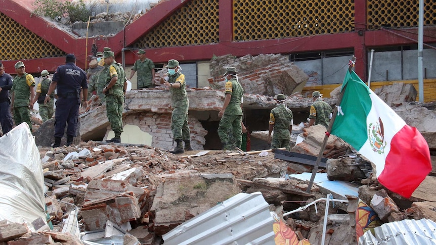 A group of soldiers stand on top of rubble with a Mexican flag next to them.