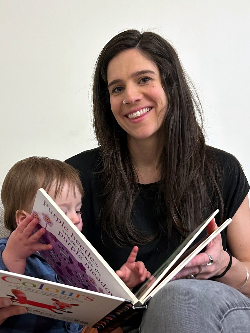 Ana Kresina smiles while reading a book to her young child. She's wearing a black t-shirt.