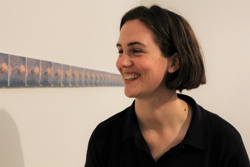 A beautiful young woman smiles looking at a sequence of 48 small oil paintings hung in a straight line