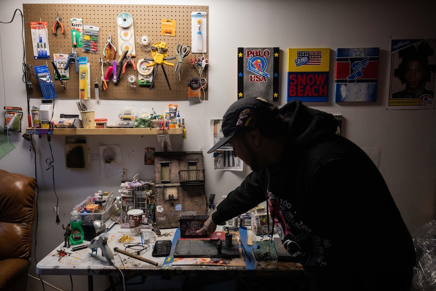 A man stands over a desk covered in tools, materials and miniature models of street scenes