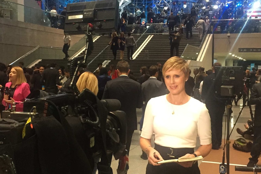 A tall blonde reporter stands before cameras in a cavernous room, decorated with American flags