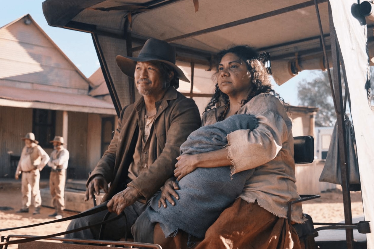 Deborah Mailman sits on a horse-drawn cart in the film Wolfram while in character. She is holding a small baby. 