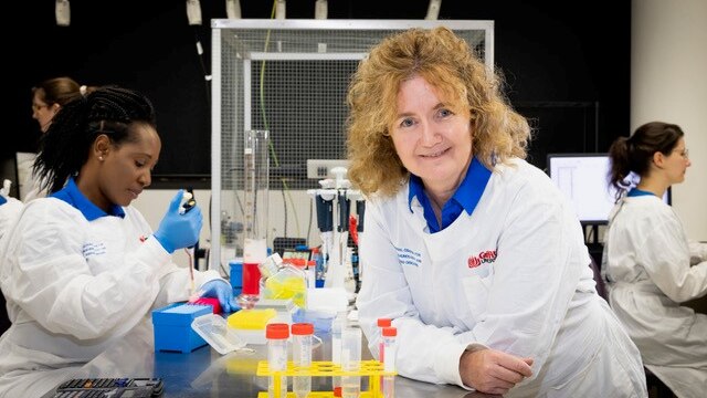 A smiling scientist with frizzy hair leans on a bench in a laboratory.