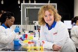 A smiling scientist with frizzy hair leans on a bench in a laboratory.