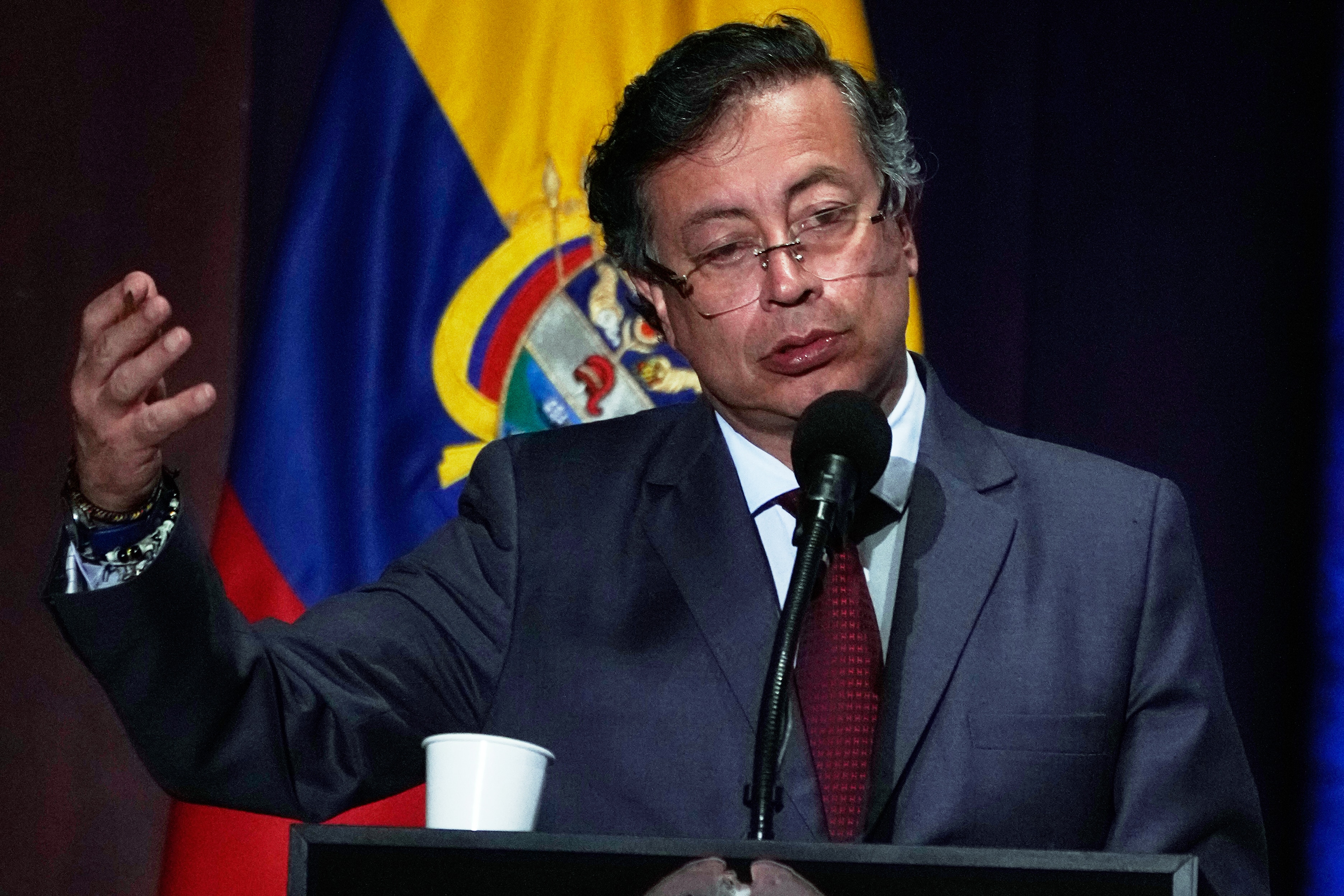 Gustavo Petro in a dark suit speaking at a lectern and gesturing with his right hand in front of a Colombian flag