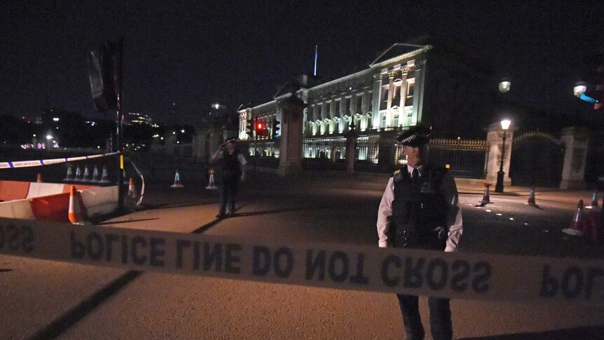 Police officer stands inside a police cordon outside Buckingham Palace. Police tape is blocking off access to the palace gate.