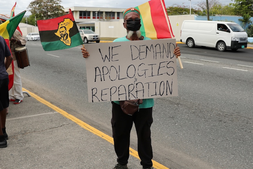 A Jamaican man holds up a placard that reads 'we demand apologies and reparation'