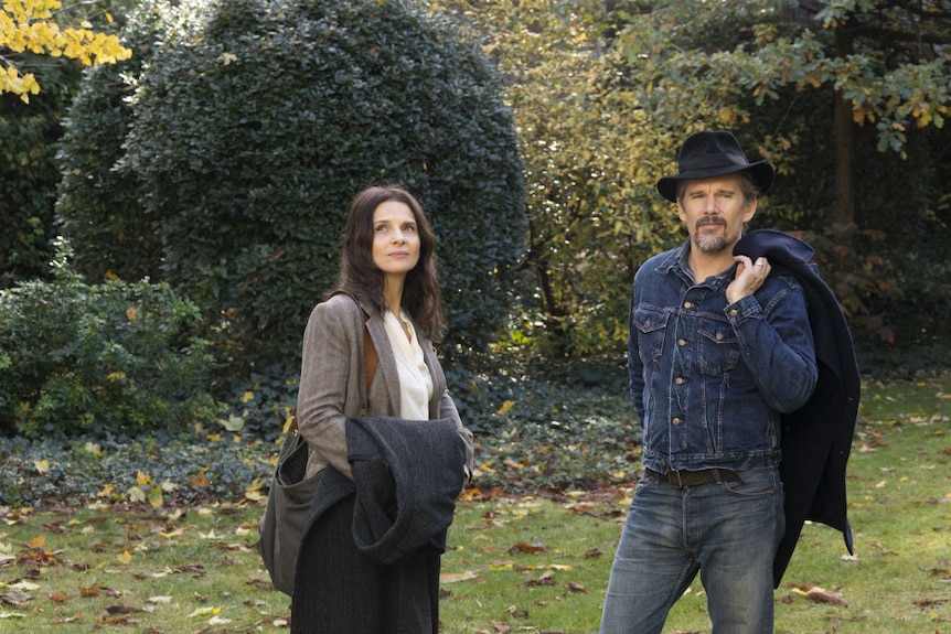 A woman in a jacket holding a coat and a man in double denim, a brimmed hat, and a coat over his shoulder in a park