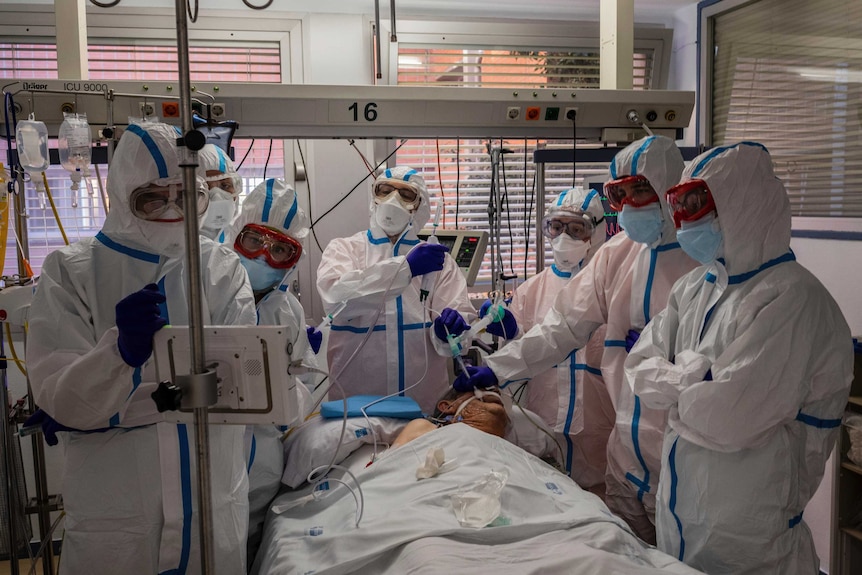 Con el EPP completo, ocho médicos se paran alrededor de la cama de un paciente anciano