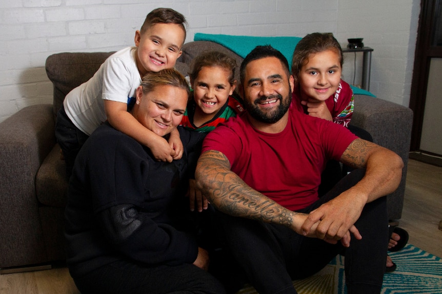 A family of five, two adults and three children, pictured sitting in front of a brown sofa.