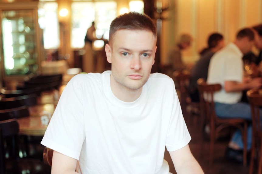 Young man with short blond hair wearing white tee shirt sits at cafe table.