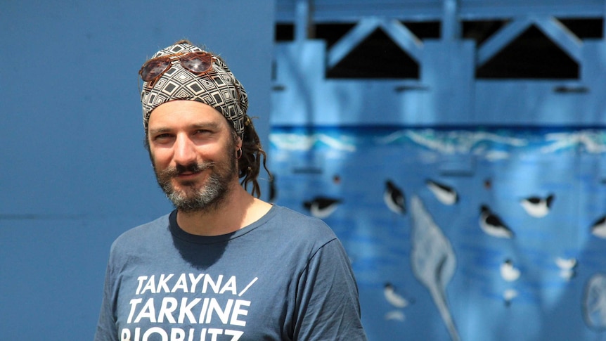 A man with a beard, wearing a blue tshirt and sunglasses on top of his head standing in front of a blue background