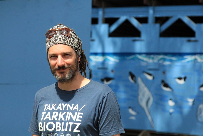 A man with a beard, wearing a blue tshirt and sunglasses on top of his head standing in front of a blue background