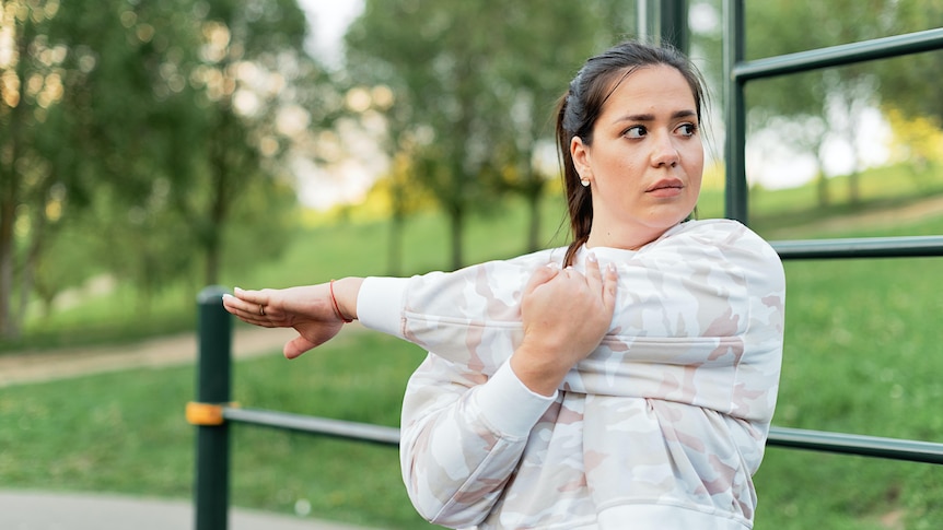 A woman stretching in a park, while looking suspiciously to the side. 