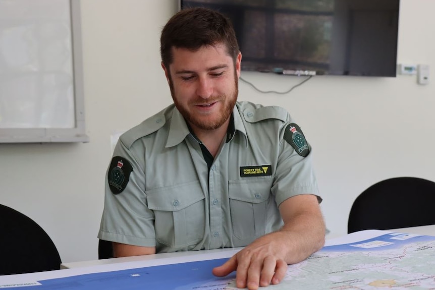 A young man in a green forest fire management uniform sits at a table in a board room and points to a map on the table.