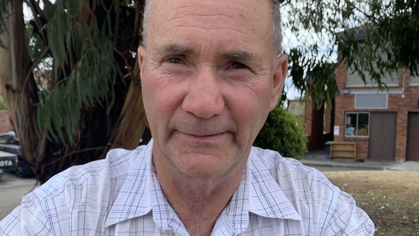 A man smiles into the camera as he sits in front of a tree