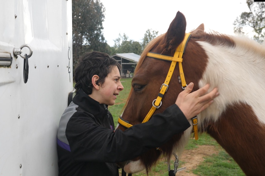 teenage boy pats his horse, which is wearing a yellow bridle, by a horse float