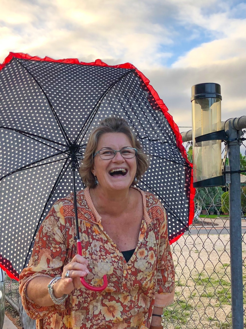 A woman laughs as she holds a spotted umbrella with red trim next to a rain gauge.