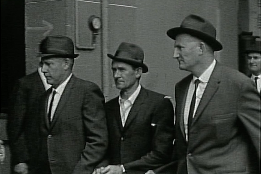 A historical black-and-white image of two police officers escorting a well-dressed criminal into a courthouse through a crowd