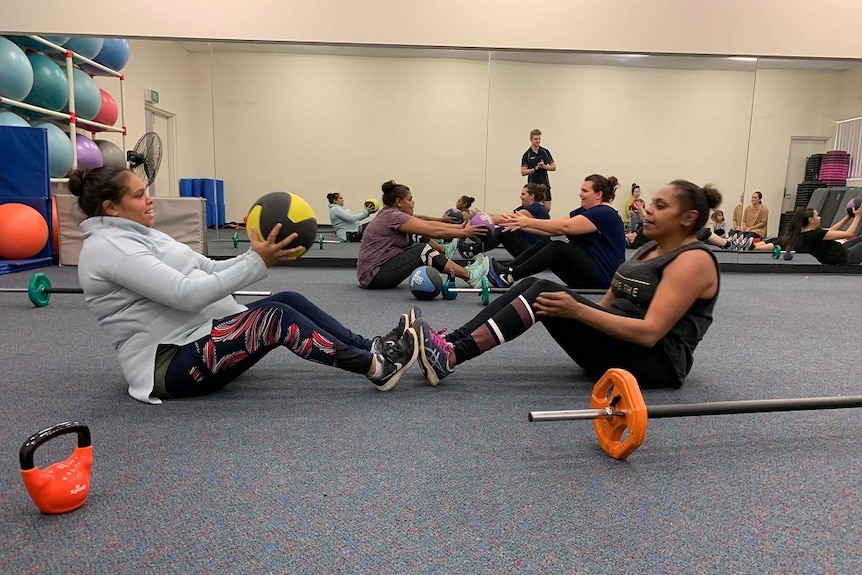 A group of women are sitting on the floor, passing medicine balls to each other. Their feet are touching.