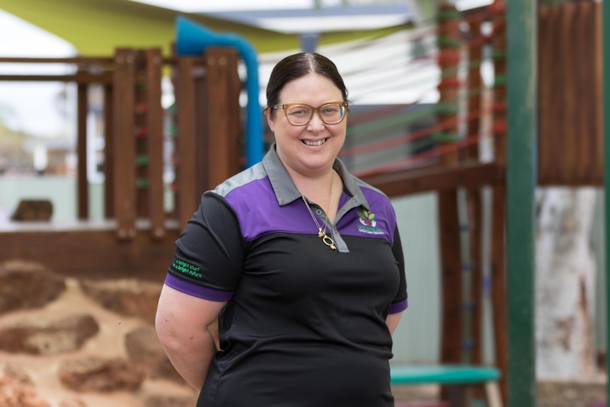 A woman in a playground at a child care centre holding her hands behind her back and smiling.  