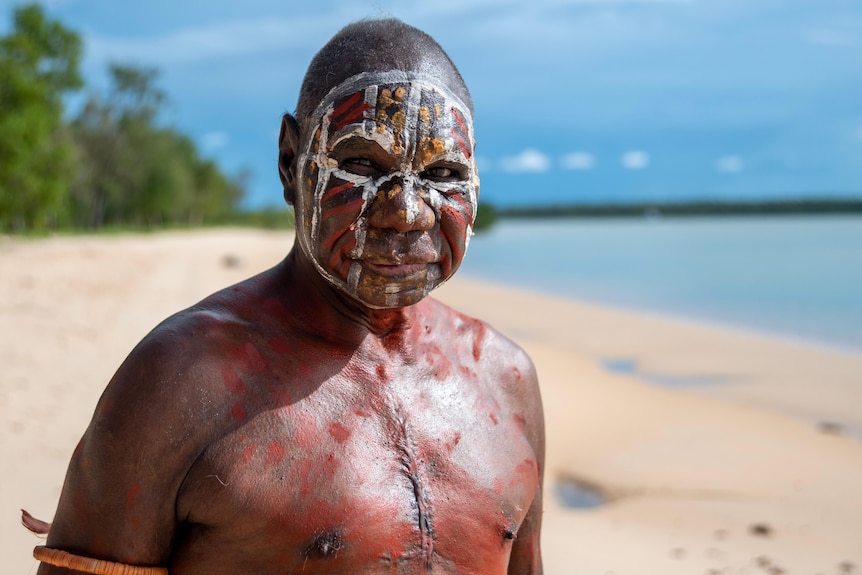 A man in traditional aboriginal painting looks directly at the camera