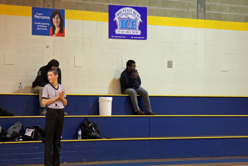 A man sits apart from the crowd, speaking into his smartphone, sitting on the bleachers of a suburban gymnasium.