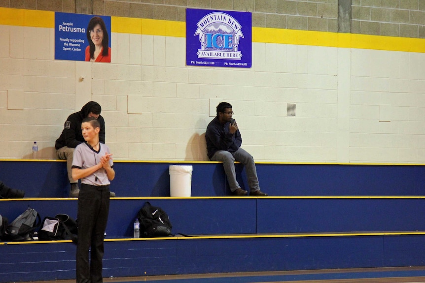 A man sits apart from the crowd, speaking into his smartphone, sitting on the bleachers of a suburban gymnasium.
