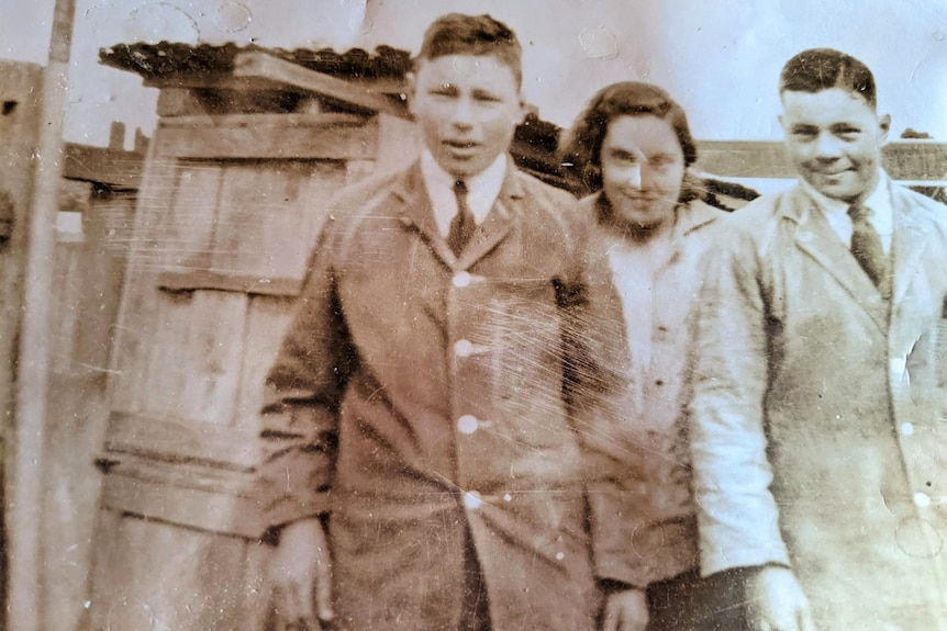 An old discoloured photo of a young man with a button down coat, his sister with dark shoulder length hair and another man.