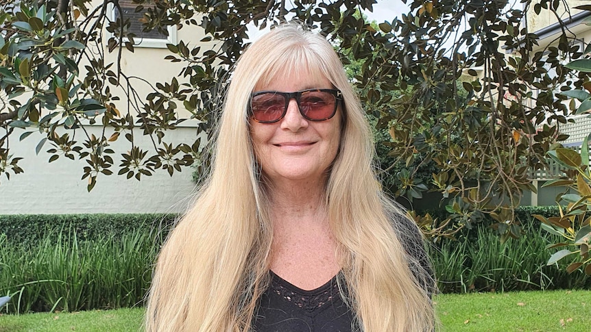 A woman with long blonde hair wears sunglasses and smiles at the camera, she stands in a small park with trees and grass.