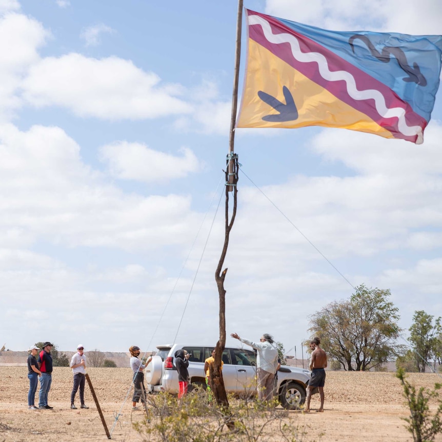A small group of people gather in a rocky, desert landscape with one car and one police car. There is a flag in the foreground.