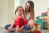 A woman with long hair and glasses smiles at a young blonde girl sitting on her lap, with building blocks on the floor.