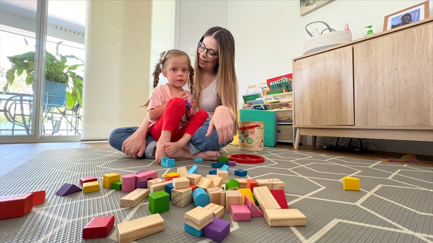 A woman with long hair and glasses smiles at a young blonde girl sitting on her lap, with building blocks on the floor.