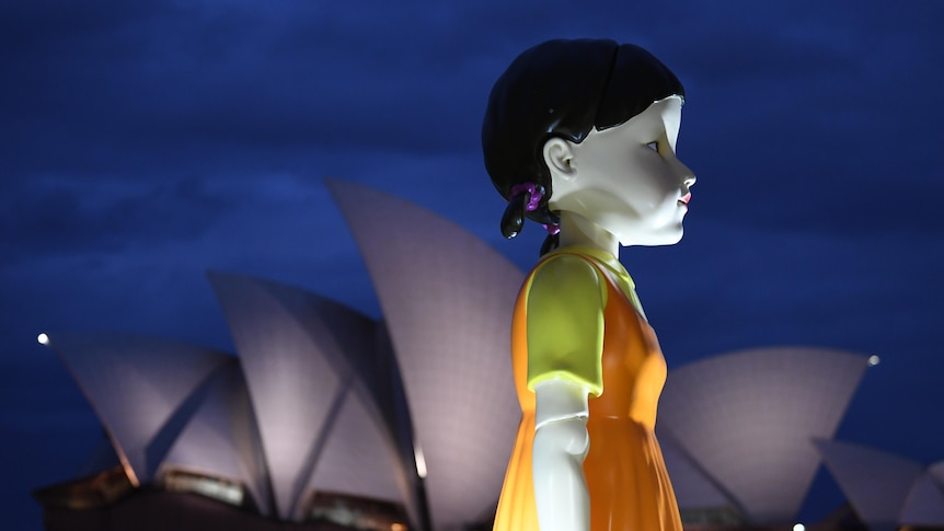 A giant pig tailed doll stands in front of the Sydney Opera House at night.