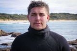 Portrait of young man with wetsuit and wet hair standing on a rock platform.
