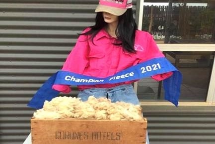 A lady scarecrow stands in front of a wool fleece holding a blue ribbon with the words Champion Fleece 2021